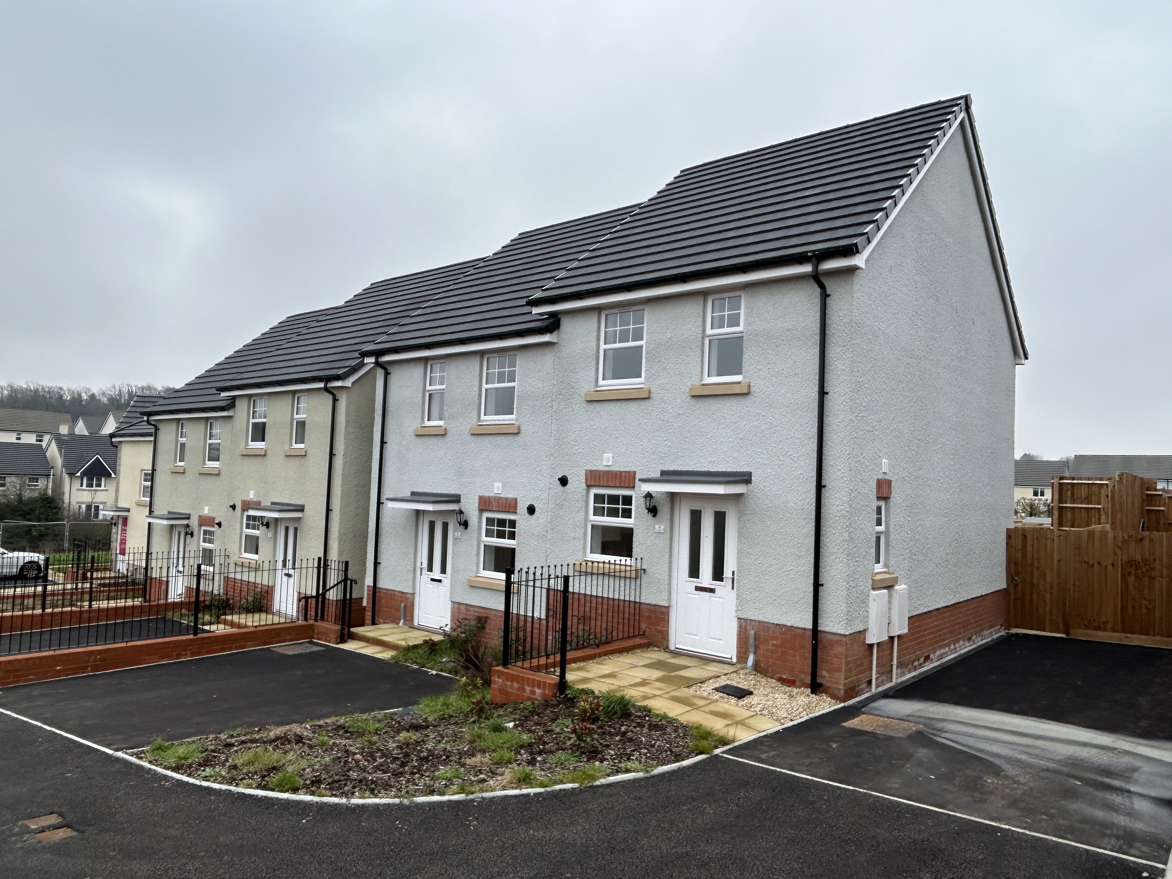 A row of newly built semi-detached houses with light-coloured rendered walls and dark grey tiled roofs. Each home has a small front garden area with low black railings, paved paths, and white front doors. The houses sit beside a freshly laid tarmac road.